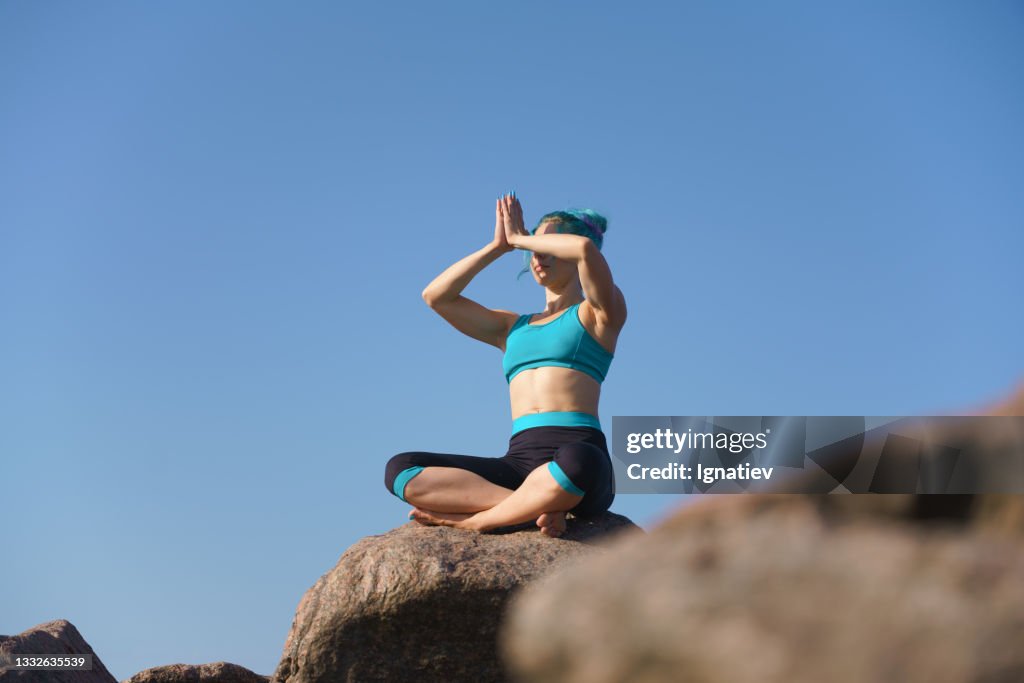 Attractive Beautiful Fit Young Woman With A Turquoise Hair In A Turquoiseblack Yoga Outfit Sits On A Stone Against The Sky With Her Hands In Front Of Her Face Palms Together High-Res Attractive Beautiful Fit Young Woman With A Turquoise Hair In A Turquoiseblack Yoga Outfit Sits On A Stone Against The Sky With Her Hands In Front Of Her Face Palms Together High-Res