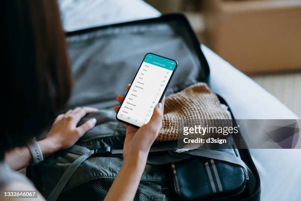 over the shoulder view of young asian woman using smartphone, checking the travel checklist while packing a suitcase on bed for a trip at home. staycation, travel and vacation concept - vacation planning stock pictures, royalty-free photos & images