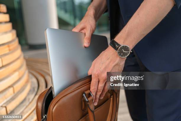 a close up of an entrepreneur  in a blue suit with a good wrist watches is standing near the wooden bech against the modern facade of a business center puts his laptop in a leather laptop bag - leather bag stock pictures, royalty-free photos & images