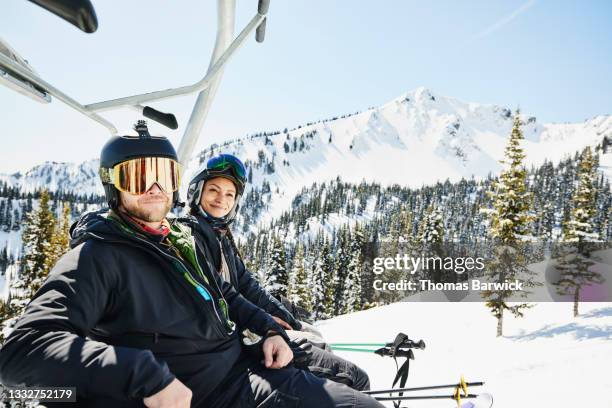 medium wide shot portrait of smiling couple riding chair lift at resort on sunny winter afternoon - skidsemester bildbanksfoton och bilder