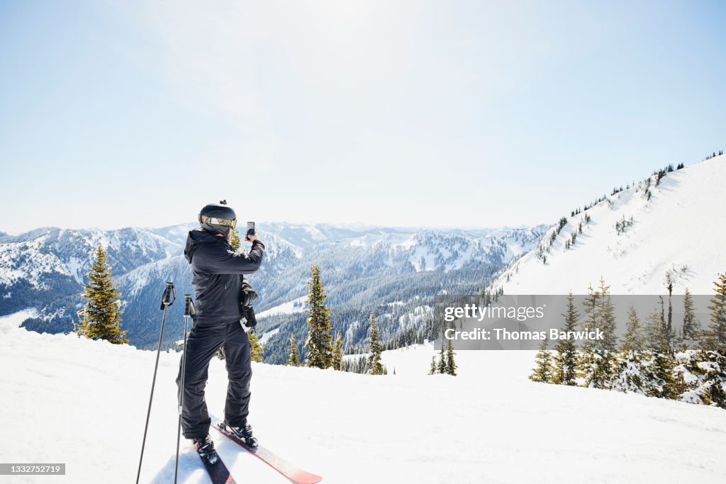 Wide shot of male skier taking photo of ski area and mountains while skiing on sunny winter morning