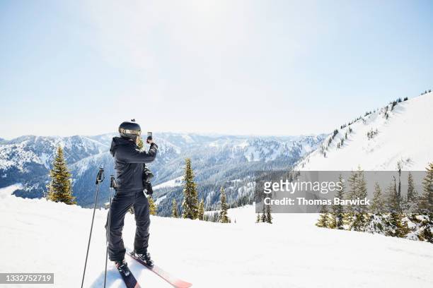 wide shot of male skier taking photo of ski area and mountains while skiing on sunny winter morning - ski pants stock-fotos und bilder