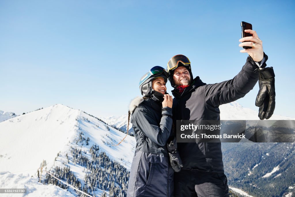 Medium wide shot of smiling couple taking selfie while standing at the top of ski resort on winter morning
