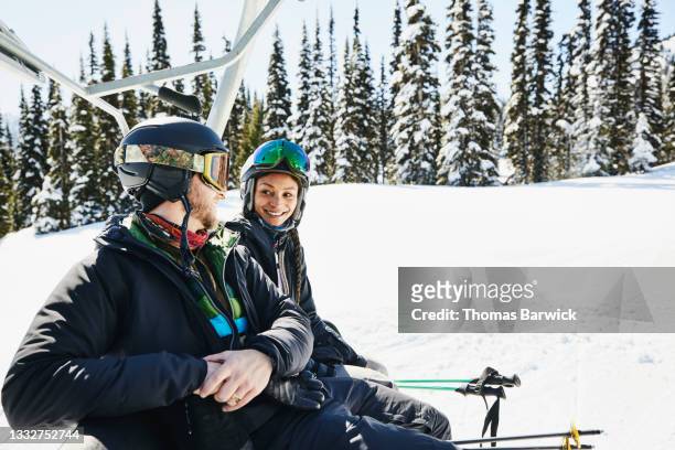 medium wide shot of smiling couple in discussion while riding chair lift at resort on sunny winter afternoon - sessellift stock-fotos und bilder
