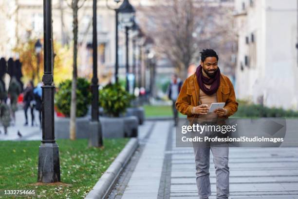a young african-american with digital tablet is drinking coffee and walking down the city district. - smart casual stock pictures, royalty-free photos & images