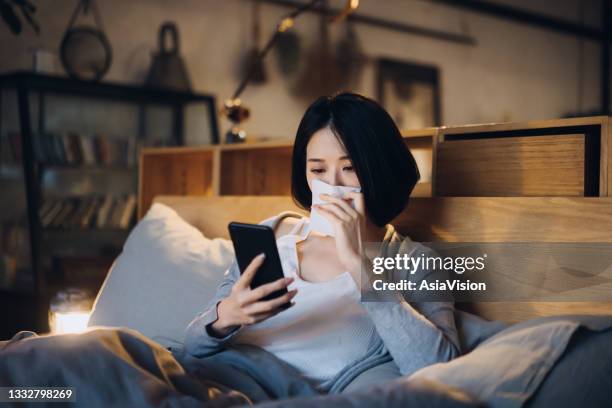 young asian woman suffering from cold and flu, blowing her nose with tissue while using smartphone on the bed and taking a rest at home - nightwear stock pictures, royalty-free photos & images