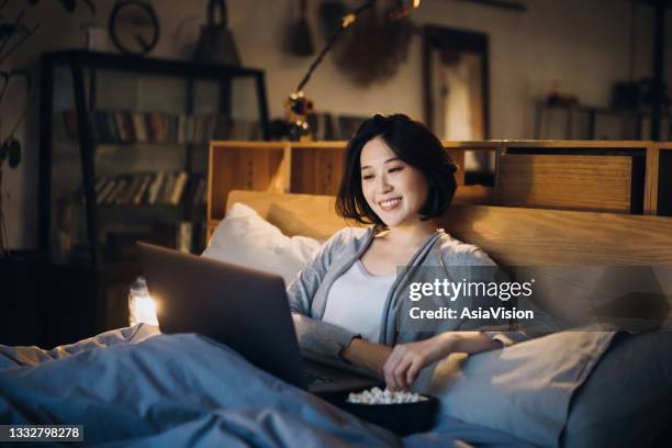 cheerful young asian woman lying on bed enjoying the weekend, watching movie on laptop and eating popcorn at home in the evening - asian watching movie stockfoto's en -beelden