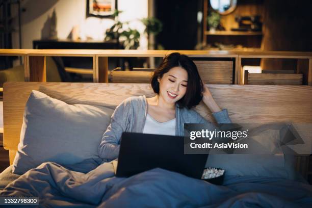 cheerful young asian woman lying on bed enjoying the weekend, watching movie on laptop and eating popcorn at home in the evening - asian cinema bildbanksfoton och bilder