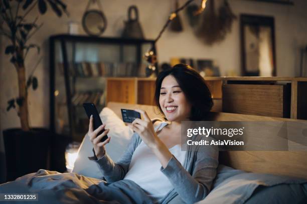 beautiful smiling young asian woman lying on bed, shopping online on her smartphone and making mobile payment with credit card at home in the evening - east asia stock pictures, royalty-free photos & images