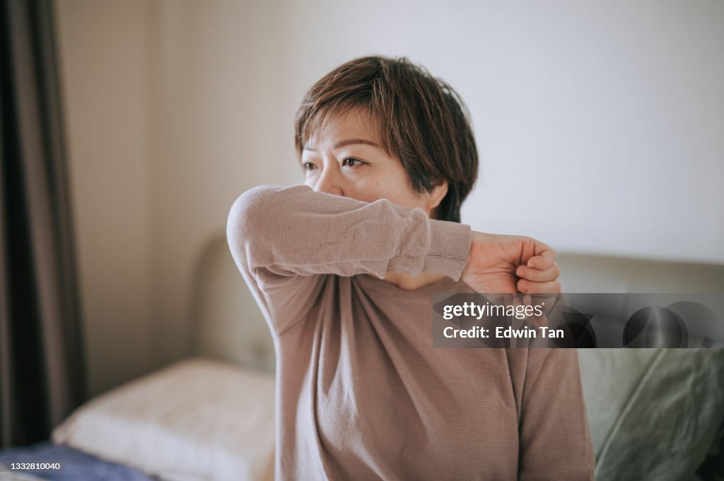 Asian chinese mid adult woman coughing into elbow not feeling well sick at home in bedroom resting