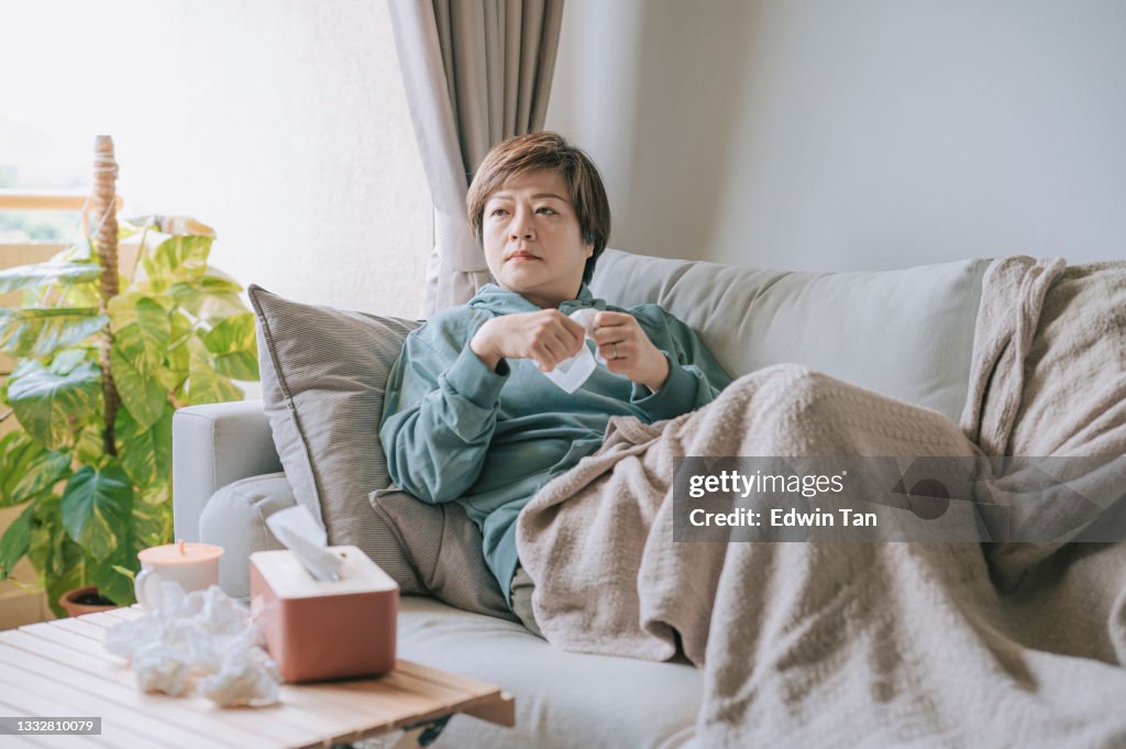 Asian chinese mid adult woman coughing with tissue paper not feeling well sick at home sitting on sofa watching tv resting