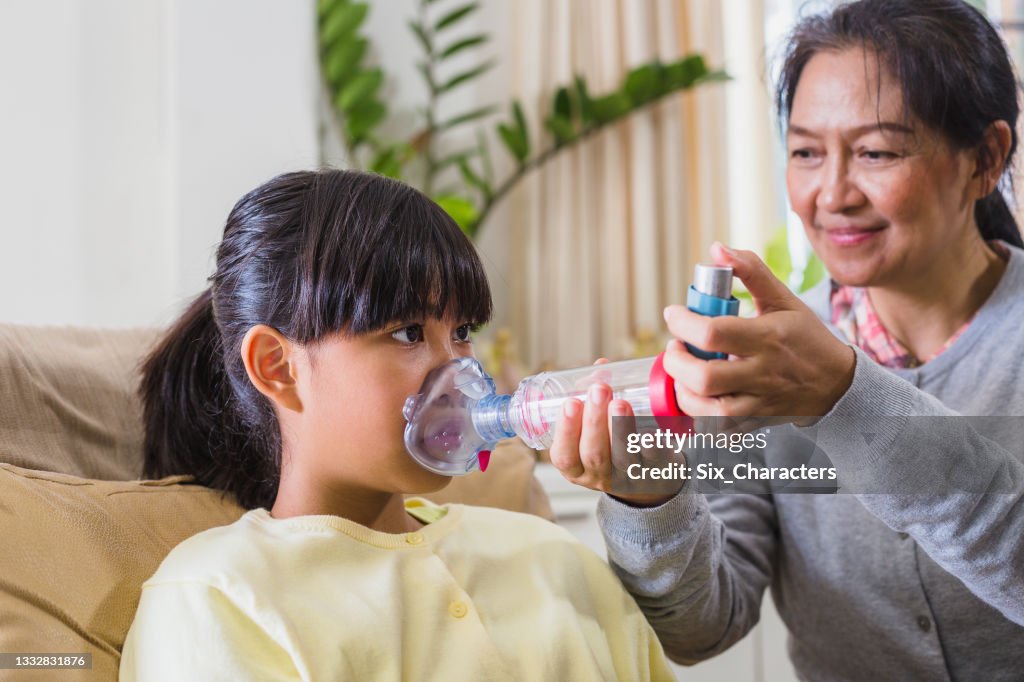 Mãe asiática ajuda filha a receber assistência com espaçador inalador de asma enquanto se instala em sofá na sala de estar em casa