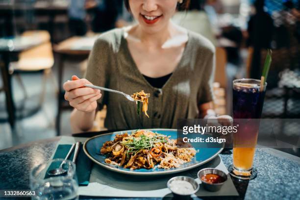 cropped shot of joyful young asian woman eating freshly served authentic malaysian-style flat rice noodle with iced fruit tea in a fusion restaurant. an iconic taste of local malaysian cuisine. asian style cuisine and food culture. eating out lifestyle - thai food stock pictures, royalty-free photos & images
