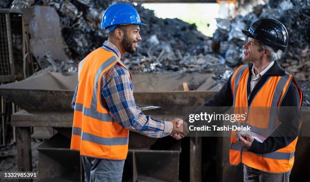 side view of two colleagues in safety reflective vests standing in factory in front of pile of scrap metal - junkyard stockfoto's en -beelden