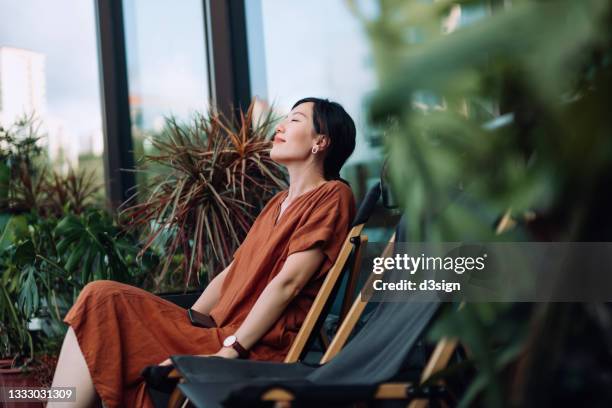 young asian woman with eyes closed enjoying fresh air while relaxing on deck chair in balcony in the morning, surrounded by beautiful houseplants. lifestyle and wellbeing concept - still leben stock-fotos und bilder