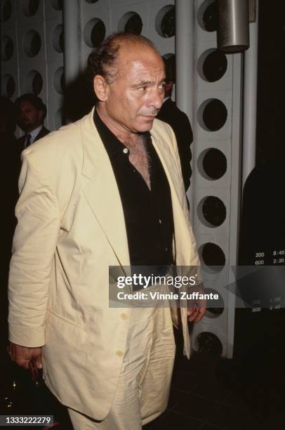 American actor Burt Young, wearing a cream-coloured lightweight suit and a black shirt open at the collar, carrying a briefcase, location...