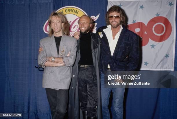 British pop group The Bee Gees pose in front of a blue curtain on which are banners for 'Coca Cola Classic' and 'HBO', at an unspecified event,...