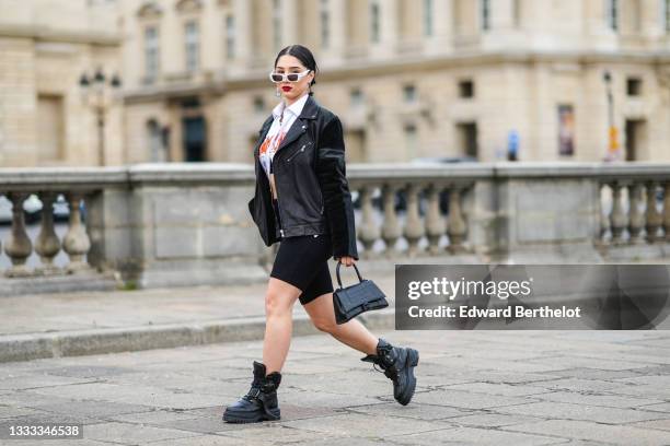 Lycia Lamini wears white sunglasses, black and white large Japanese character pendant earrings, a silver and rhinestones chain necklace, a silver...