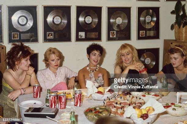 American New Wave band The Go-Go's seated at a table on which is a plate of sandwiches, bowls of food and cans of Coca Cola, with presentation discs...