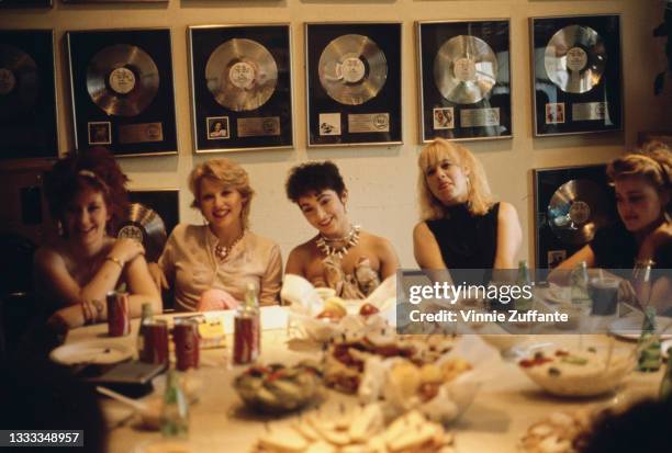 American New Wave band The Go-Go's seated at a table on which is a plate of sandwiches, bowls of food and cans of Coca Cola, with presentation discs...