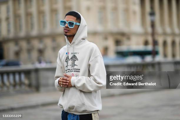 Zayd Cus wears pale blue sunglasses, a beige hoodie oversized sweater with logo and slogan "Police Nationale", blue faded denim ripped jeans pants...