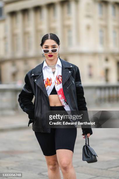 Lycia Lamini wears white sunglasses, black and white large Japanese character pendant earrings, a silver and rhinestones chain necklace, a silver...