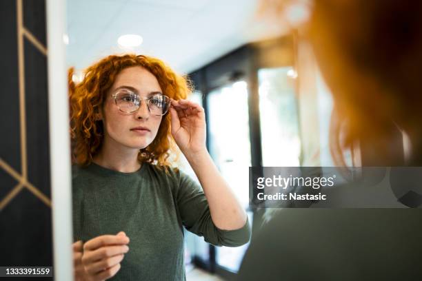 mujer joven probando gafas en tienda óptica mirando al espejo - lente fotografías e imágenes de stock