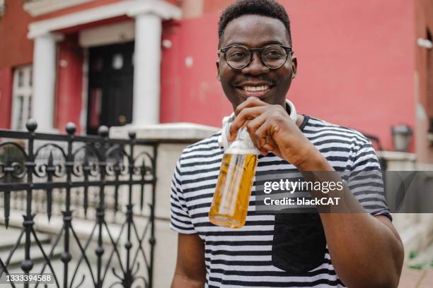 young african-american man is drinking beer on the street - man sipping beer smiling stockfoto's en -beelden