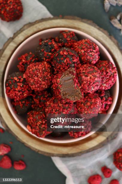 close-up image of homemade, individual chocolate truffle sweets with ganache centre, piled in pink bowl on a wooden board, luxury chocolate balls coated in dehydrated raspberry pieces, pink muslin, missing bite, dark grey background, elevated view - milk chocolate truffles stock pictures, royalty-free photos & images