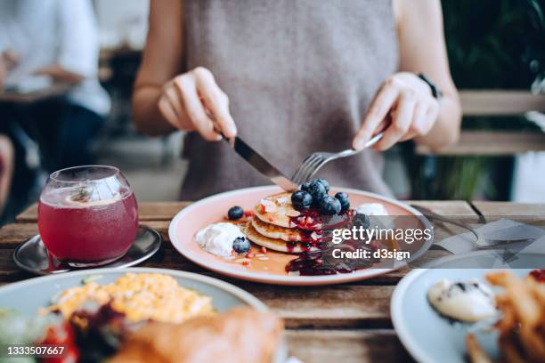 close up of young woman sitting at dining table eating pancakes with blueberries and whipped cream in cafe, with english breakfast and french fries served on the dining table. eating out lifestyle - blueberry pancakes stock-fotos und bilder