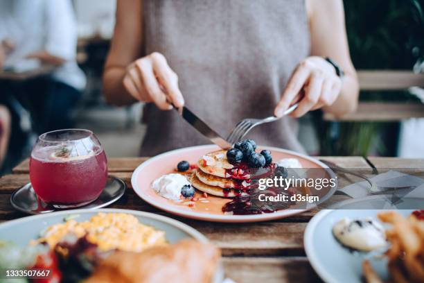 close up of young woman sitting at dining table eating pancakes with blueberries and whipped cream in cafe, with english breakfast and french fries served on the dining table. eating out lifestyle - dessert stockfoto's en -beelden