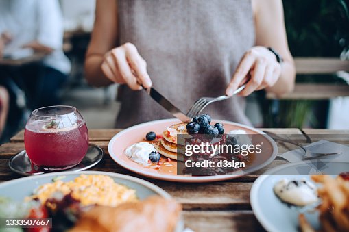 Close up of young woman sitting at dining table eating pancakes with blueberries and whipped cream in cafe, with English breakfast and french fries served on the dining table. Eating out lifestyle Close up of young woman sitting at dining table eating pancakes with blueberries and whipped cream in cafe, with English breakfast and french fries served on the dining table. Eating out lifestyle