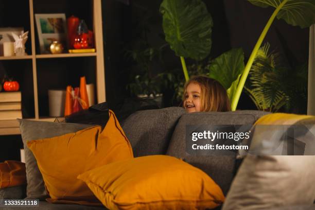 cheerful little girl playing hide and seek and peeking behind a sofa - kid hide and seek stock pictures, royalty-free photos & images