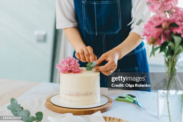 dark-haired woman wearing apron decorate сake with fresh peonies and leaves in the kitchen - decorating a cake stock pictures, royalty-free photos & images