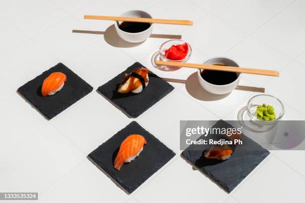 salmon and unagi sushi, soy sauce, bamboo sticks, wasabi, ginger on white tiled table and slate stone coasters. composition with hard light. - wasabi photos et images de collection