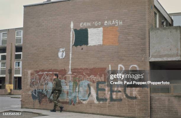 British Army soldier on patrol walks past Irish Republican Army and Irish tricolour graffiti painted on an end wall of Unity Flats in the New Lodge...