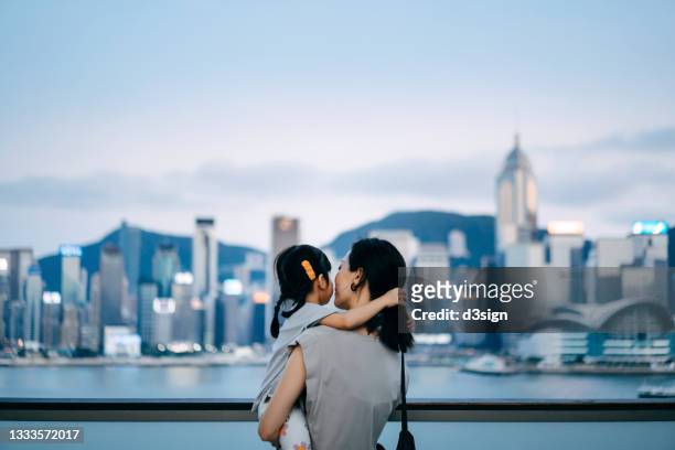 rear view of young asian mother embracing little daughter in arms, spending intimate bonding time over the urban terrace in downtown district against urban city skyline and the promenade at sunset - expectations foto e immagini stock