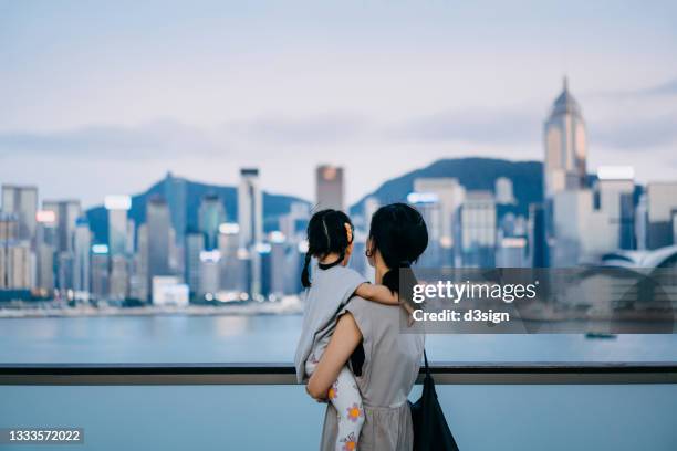 rear view of young asian mother embracing little daughter in arms, spending intimate bonding time over the urban terrace in downtown district against urban city skyline and the promenade at sunset - looking over balcony stock pictures, royalty-free photos & images