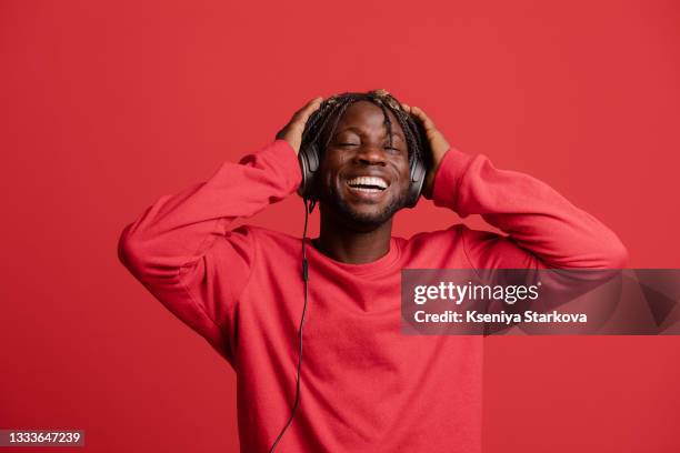 black man with short pigtails in his hair on a red background in the studio in a red jacket listens to music in headphones holds headphones with his hands smiling with closed eyes - red headphones stock pictures, royalty-free photos & images