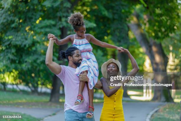 african-american parents is carrying a daughter with curly hair on shoulders, walking in the public park, and enjoying in wonderful summer afternoon. - international day eight imagens e fotografias de stock