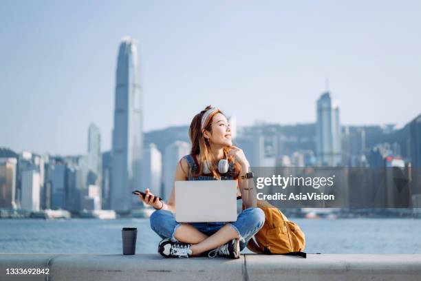 fröhliche junge asiatin sitzt im schneidersitz an der promenade, vor der skyline der stadt. sie trägt kopfhörer um den hals, benutzt ein smartphone und arbeitet am laptop, mit einer kaffeetasse an ihrer seite. mit einem lächeln wegschauen. lifestyle un - headphone man on neck stock-fotos und bilder