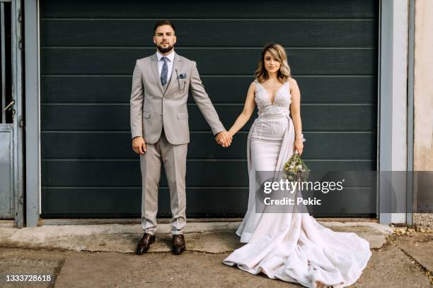 junges hochzeitspaar posiert vor garagentoren - bride and groom looking at camera stock-fotos und bilder