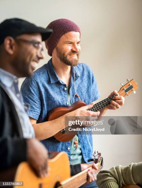 smiling man playing ukulele during a band rehearsal with friends - ukelele stock pictures, royalty-free photos & images