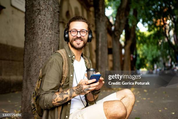 young modern man at the city street enjoying the cup of coffee while listening music on headphones, using mobile phone - persoon luisteren muziek oortjes stockfoto's en -beelden