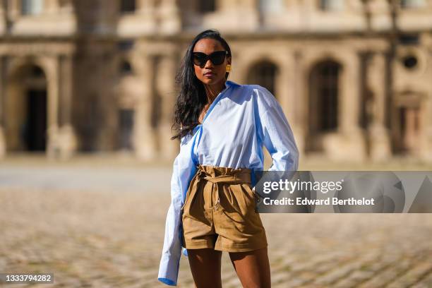 Emilie Joseph @in_fashionwetrust wears black sunglasses, an oversized off-shoulder button-down shirt in baby blue shirt, brown high waisted shorts,...