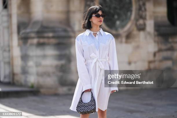 Hélène Guillaumes wears black and silver sunglasses, silver large earrings, a white pearls necklace, a white with white and blue striped yoke long...