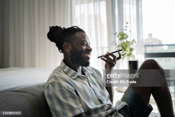 homem sentado no sofá enviando mensagem de áudio no smartphone em casa - sending - fotografias e filmes do acervo