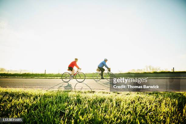 wide shot of senior male friends on sunrise bike ride on rural road - sunny side fotografías e imágenes de stock