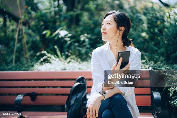 confident young asian businesswoman using smartphone, looking up while sitting on the bench in urban office park, against sunlight and green plants. taking a break and enjoying some fresh air in the middle of a work day - woman smartphone nature stockfoto's en -beelden