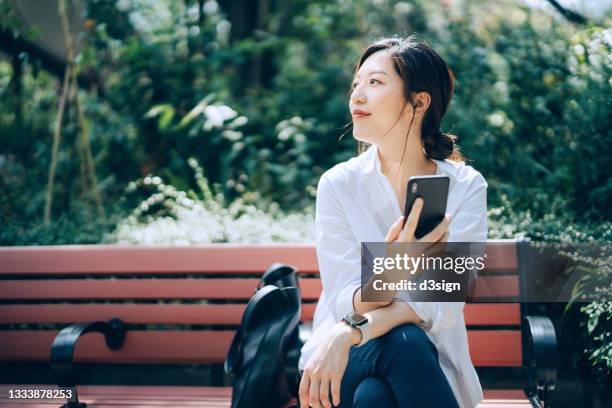 confident young asian businesswoman using smartphone, looking up while sitting on the bench in urban office park, against sunlight and green plants. taking a break and enjoying some fresh air in the middle of a work day - creative people outside imagens e fotografias de stock